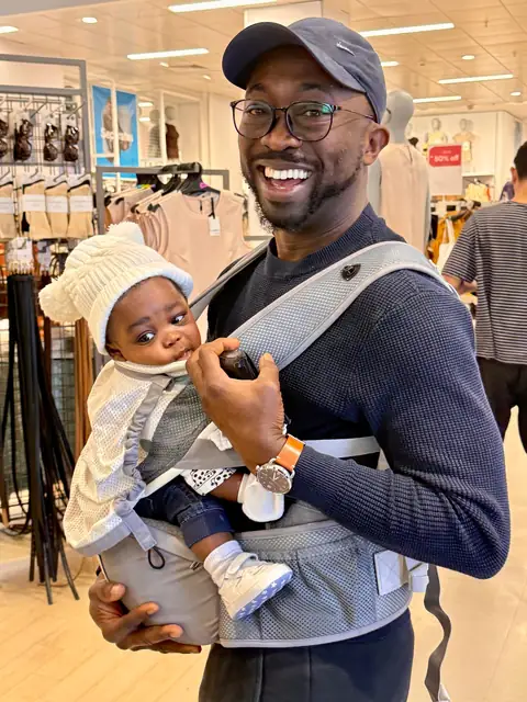 Oluseyi laughing, carrying his baby in a sling